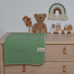 Bild mit der Decke Kinding in der Farbe Kiefernnadel. Decke liegt auf einer Kommode neben einem Teddybär und anderem Spielzeug